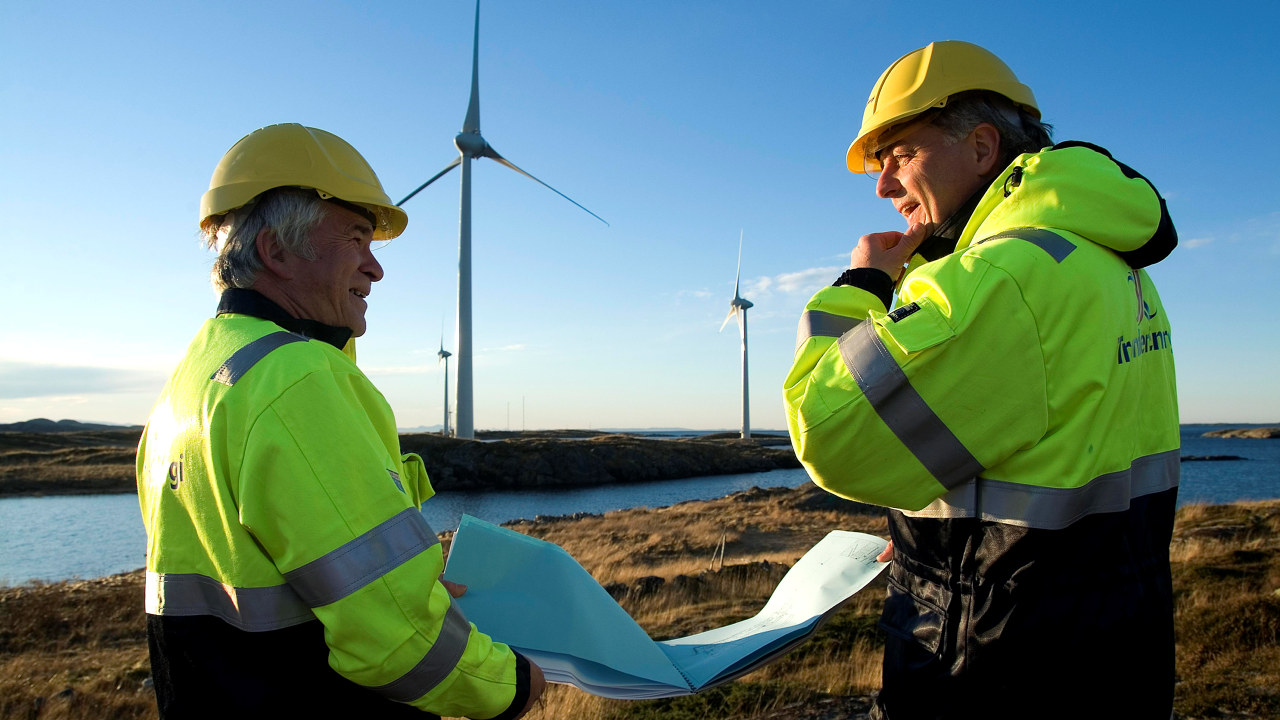 Bilde av to menn med hjelm og vernetøy som snakker sammen over et kart foran vindmøller.