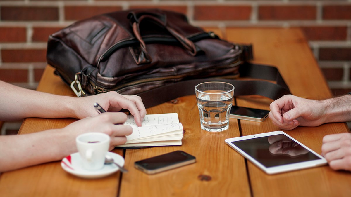 Bilde av et bord meg en bag, vannglass, kaffekopp og hender som noterer i en blokk ved siden av en mobil og et nettbrett
