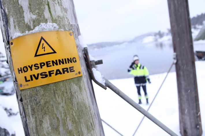 Advarselen høyspenning livsfare er festet på en kraftmast i tre og en mann i verneutstyr står i bakgrunnen. Foto