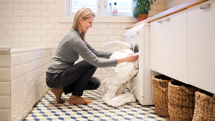 Dame med langt lyst hår sitter på huk foran en vaskemaskin med sengetøy.