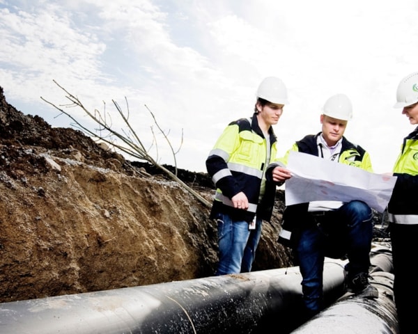 Tre mennesker står med en plantegning ved siden av fjernvarmerør. Foto