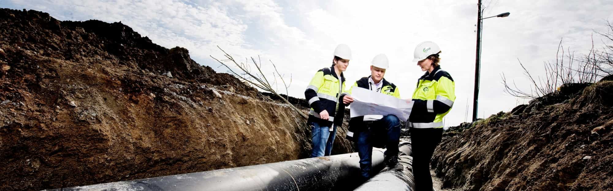 Tre mennesker står med en plantegning ved siden av fjernvarmerør. Foto